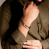 Person wearing a green shirt and beige pants with a watch on a neutral background